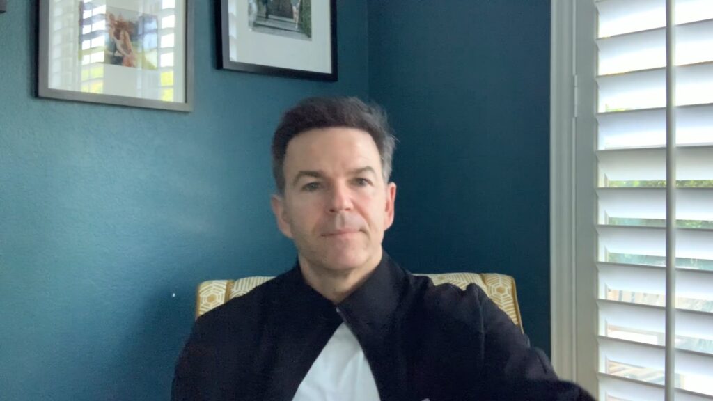 A man in a black shirt sitting on a chair against a blue wall.
